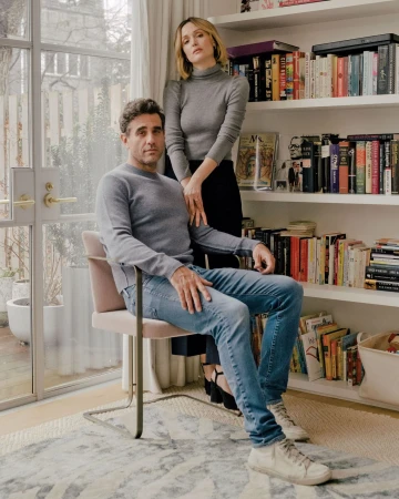 Indoor portrait of a Caucasian couple; a man sits in a chair, wearing jeans and a grey sweater; a woman stands next to him.