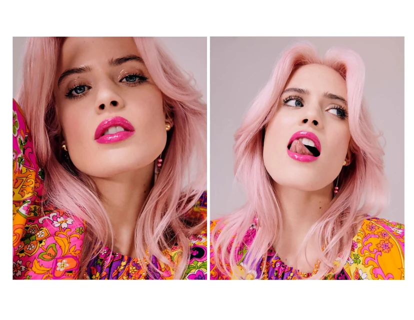 Close-up portrait of a fair-skinned woman with pink hair and bright pink lipstick, wearing a colorful patterned top in a studio shot.