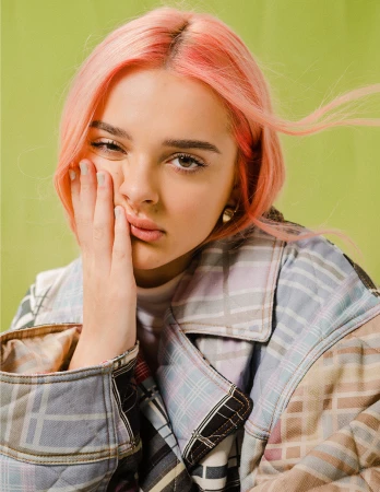 Portrait of a young woman with pink hair and a plaid quilted jacket touching her cheek against a green backdrop.