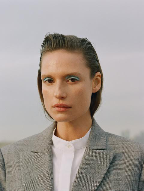 Close-up portrait of an adult woman with slicked-back brown hair, wearing a grey plaid blazer and bright blue eye shadow.