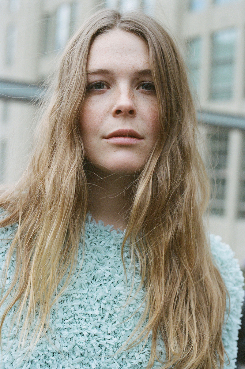 Close-up portrait of a young fair-skinned woman with wavy blonde hair wearing a blue sweater, set against an urban background.