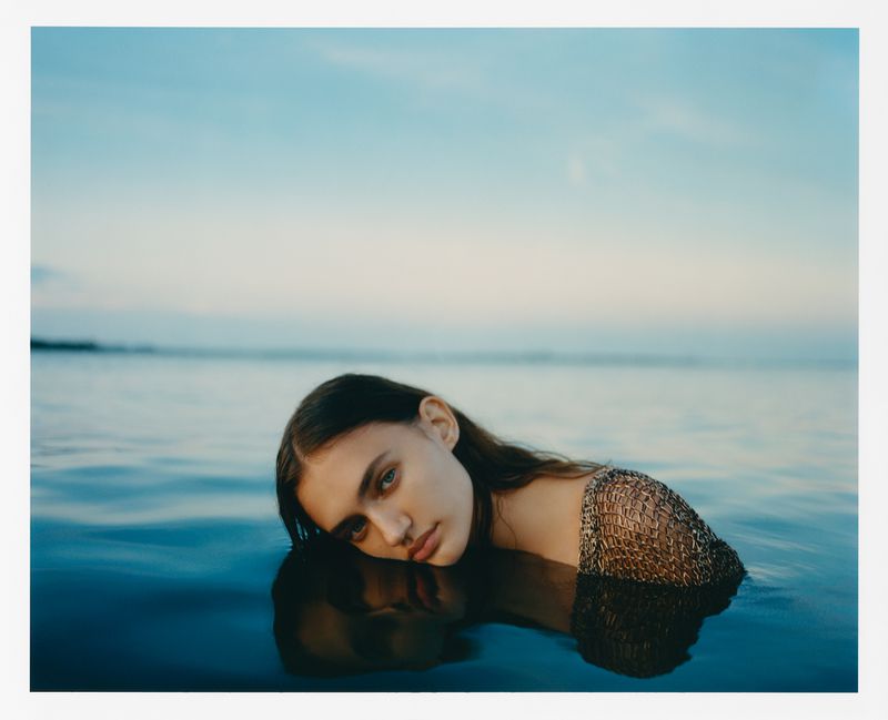 Woman in water wearing metallic mesh top, resting head on surface, open eyes, soft light, clear sky reflected on rippling water.