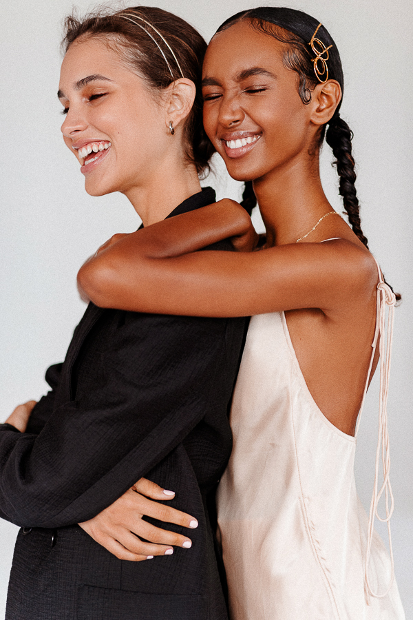 Two young women embrace closely, smiling with joy. One wears a black blazer and the other a light pink dress in a bright studio setting.
