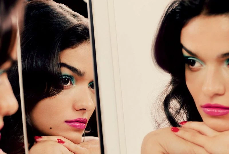 Brunette woman gazes into a mirror, showcasing vivid turquoise eyeshadow, striking pink lipstick, and a close-up, eye-level perspective.