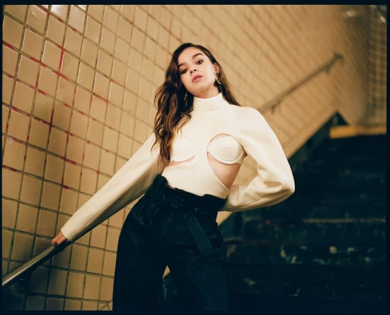 Hailee Steinfeld posing on tiled stairs wearing a white cut-out top and black trousers with hair styled by Clara Leonard