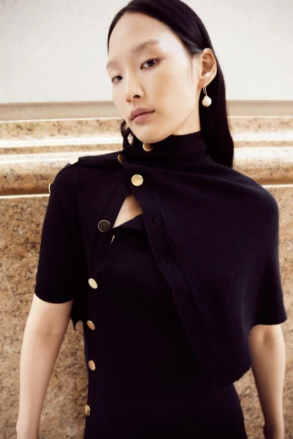 Elegant Asian woman wearing a stylish black dress with gold button accents, accessorized with pearl earrings, set against a beige backdrop.