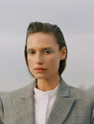 Close-up portrait of a woman with slicked-back hair, blue eyeliner, and a grey plaid blazer over a white shirt, styled by Clara Leonard.