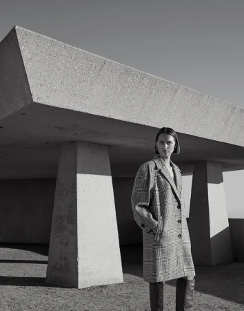 Monochrome portrait of a woman wearing a patterned trench coat and tall boots beneath a large concrete structure.