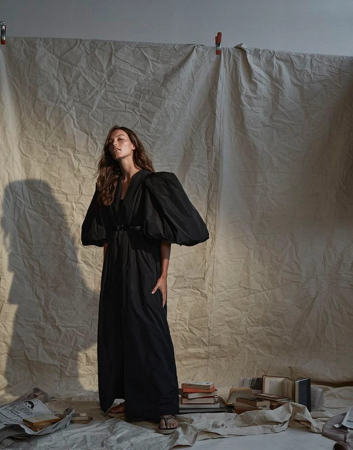 Model wearing a long black dress with voluminous puff sleeves standing against a creased backdrop.
