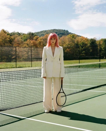 A woman with pink hair wearing a cream suit holding a tennis racket on a court with a net.