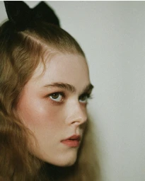 Close-up portrait of a woman with wavy hair and a black hair bow against a plain background