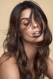Close-up portrait of a woman with dark brown wavy hair, soft lighting, and subtle hoop earrings against a warm beige background.