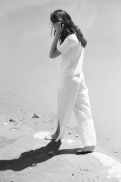Woman with long dark hair wearing a white top and pants walking barefoot on a sandy beach.