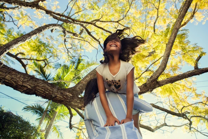 A young girl smiling with hair flying in the wind while being held up by someone beneath a large tree.