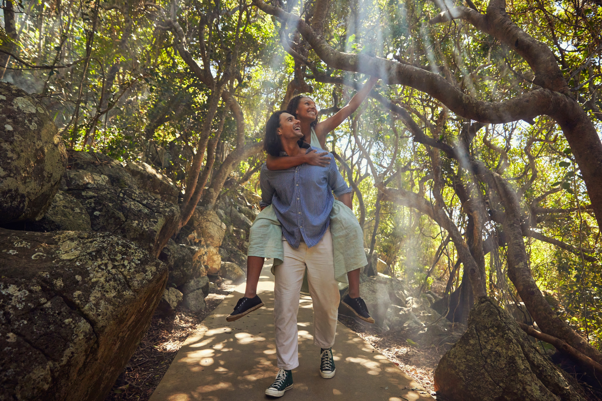 Man giving a woman a piggyback ride along a sunlit forest path framed by mossy rocks and sprawling tree branches.