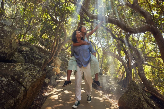 Man giving a woman a piggyback ride along a sunlit forest path framed by mossy rocks and sprawling tree branches.