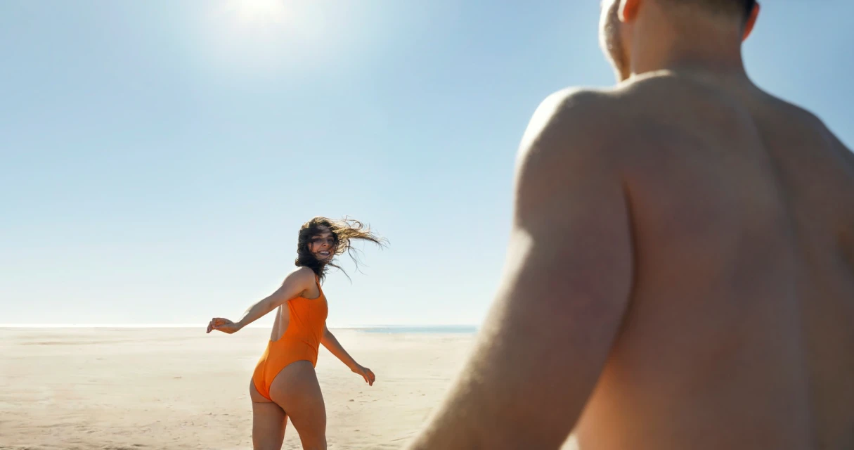 Woman in orange swimsuit runs on the beach toward the camera, pursued by a shirtless person on a sunny day with a clear blue sky.