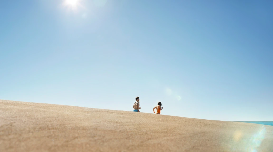 Two people running on a sand dune under a bright blue sky with the ocean visible in the distance on a sunny day.