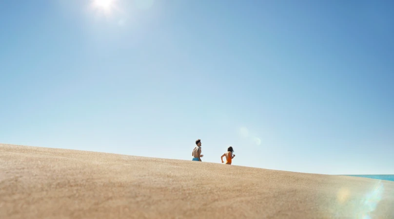 Two people running on a sand dune under a bright blue sky with the ocean visible in the distance on a sunny day.