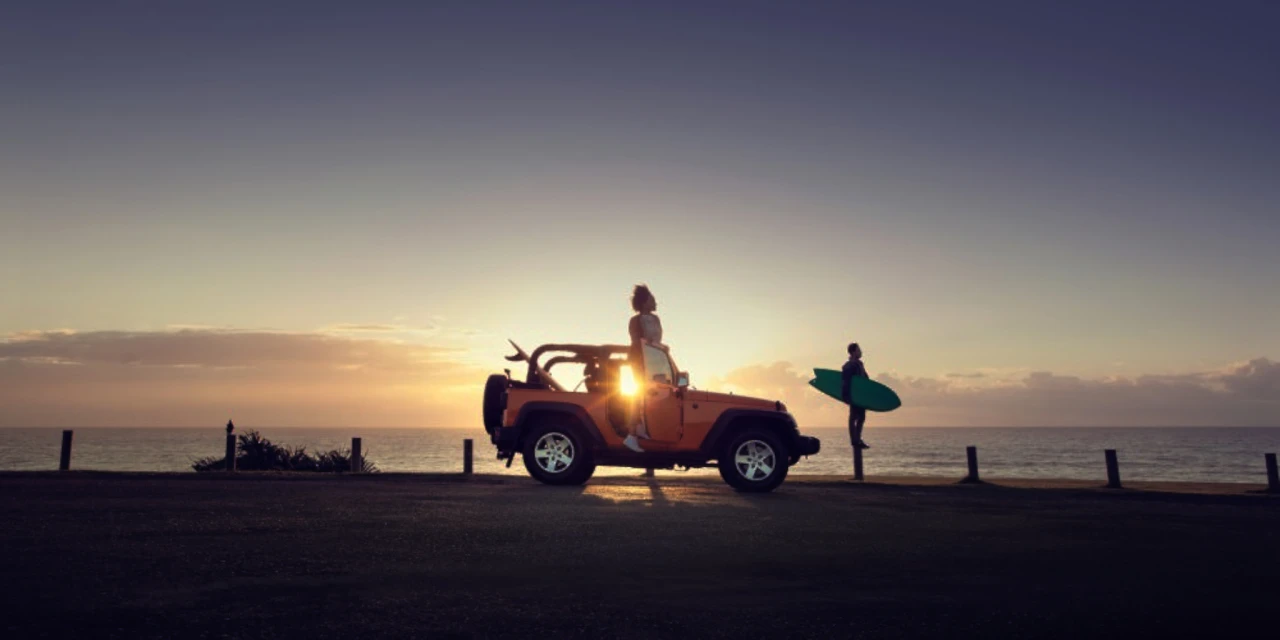 Woman sits on a yellow Jeep at sunset with a man holding a surfboard nearby against a sea and sky backdrop filled with warm sunlight.
