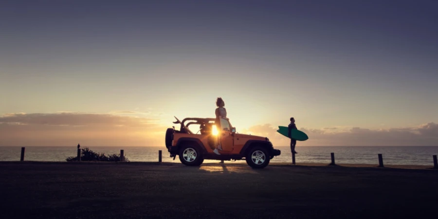 Woman sits on a yellow Jeep at sunset with a man holding a surfboard nearby against a sea and sky backdrop filled with warm sunlight.