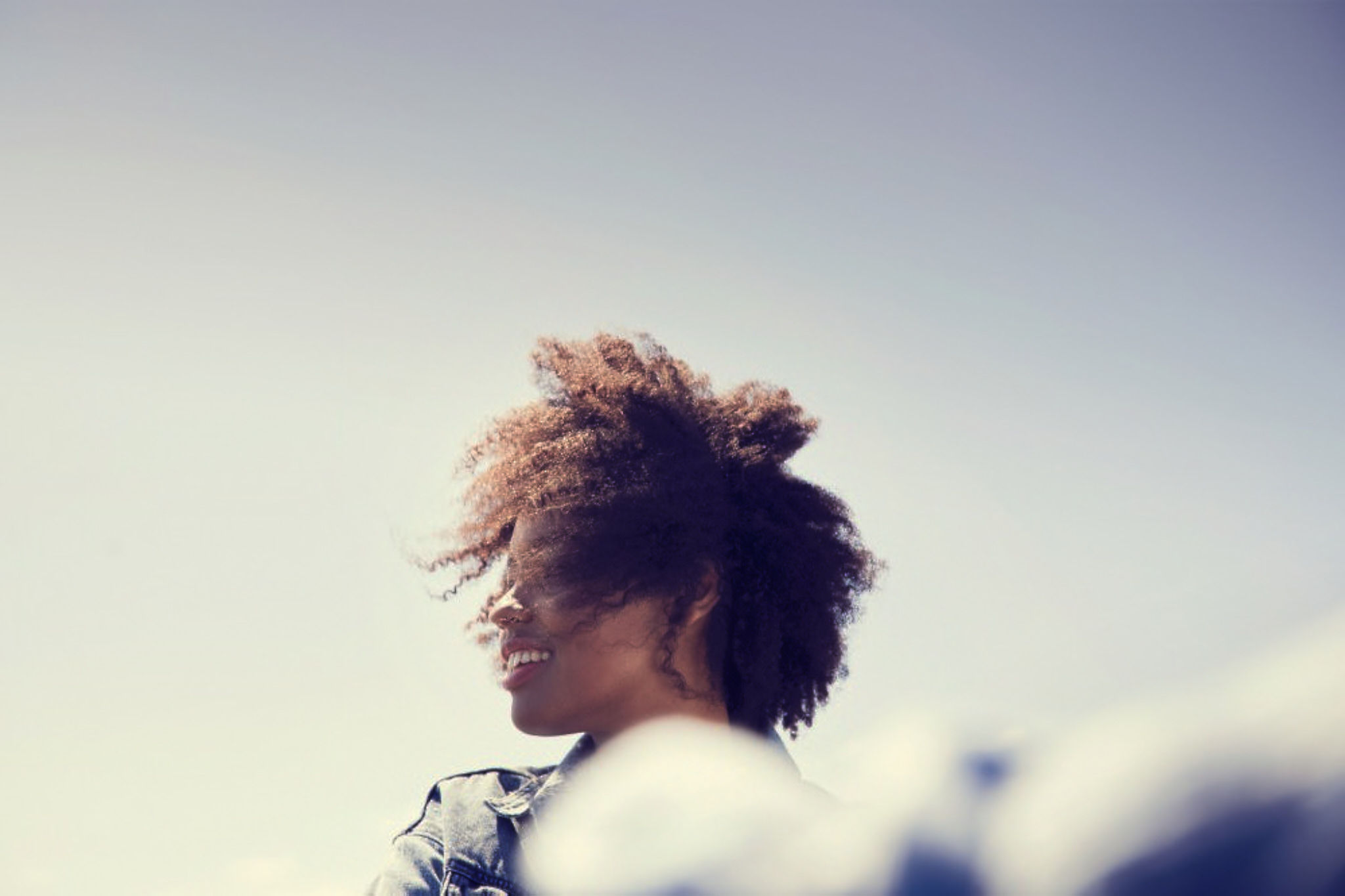 Profile of a smiling black woman with curly hair against a clear blue sky by Josh Kelly