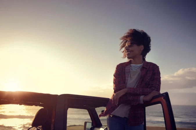 Smiling woman leaning on a car door overlooking the ocean at sunset, photographed by Josh Kelly in Sydney.