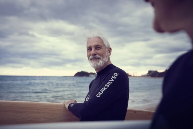 Elderly man with white hair and beard wearing black wetsuit holding surfboard near ocean by Josh Kelly