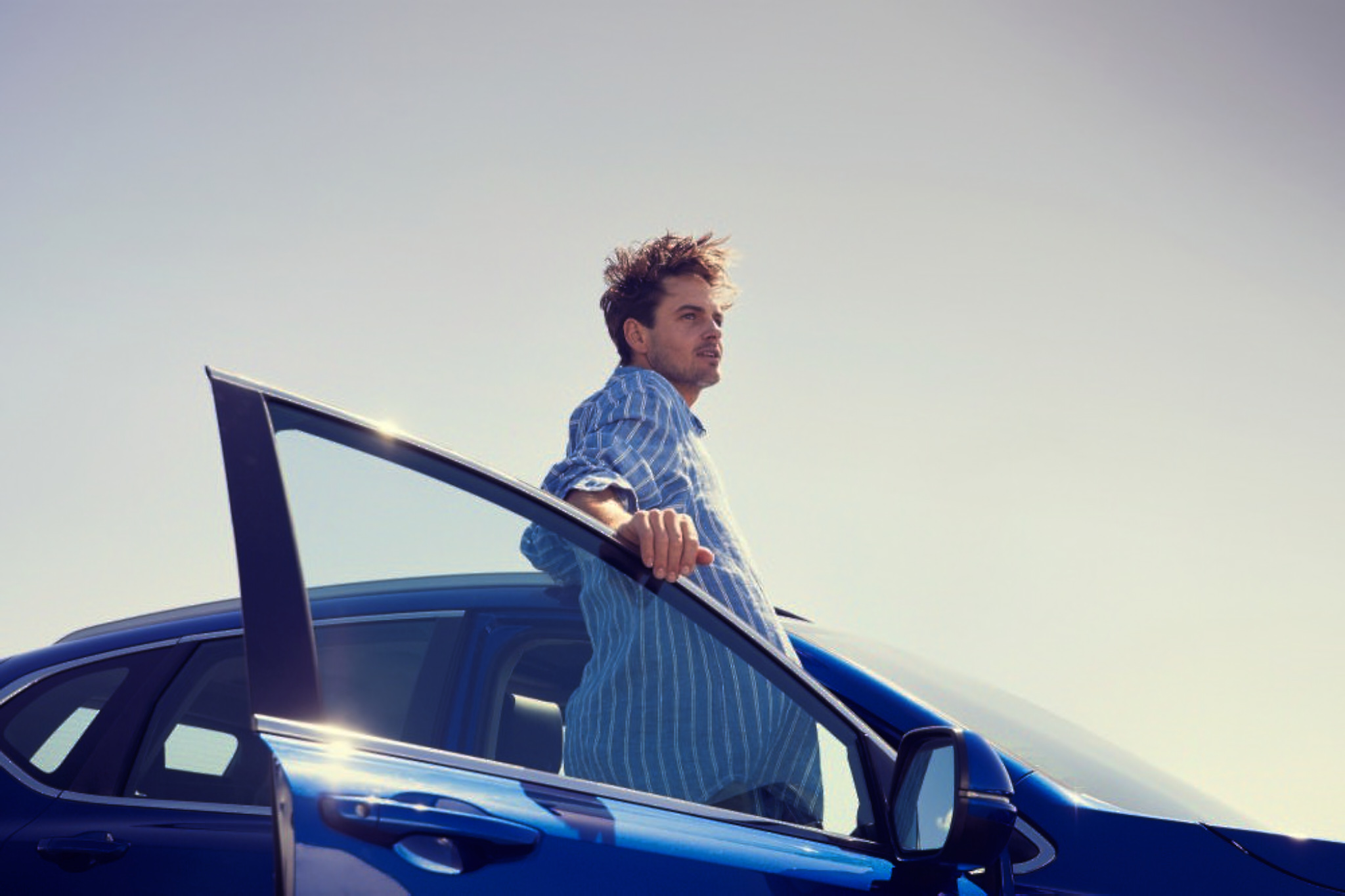 Adult male in blue shirt poses with a blue modern car with its door open under a light blue sky on a sunny day outdoors.
