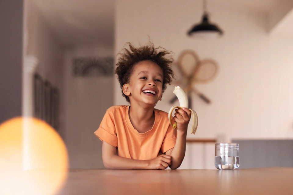 Smiling African American child sits at a wooden table holding a peeled banana, with a glass of water and a bright indoor setting.