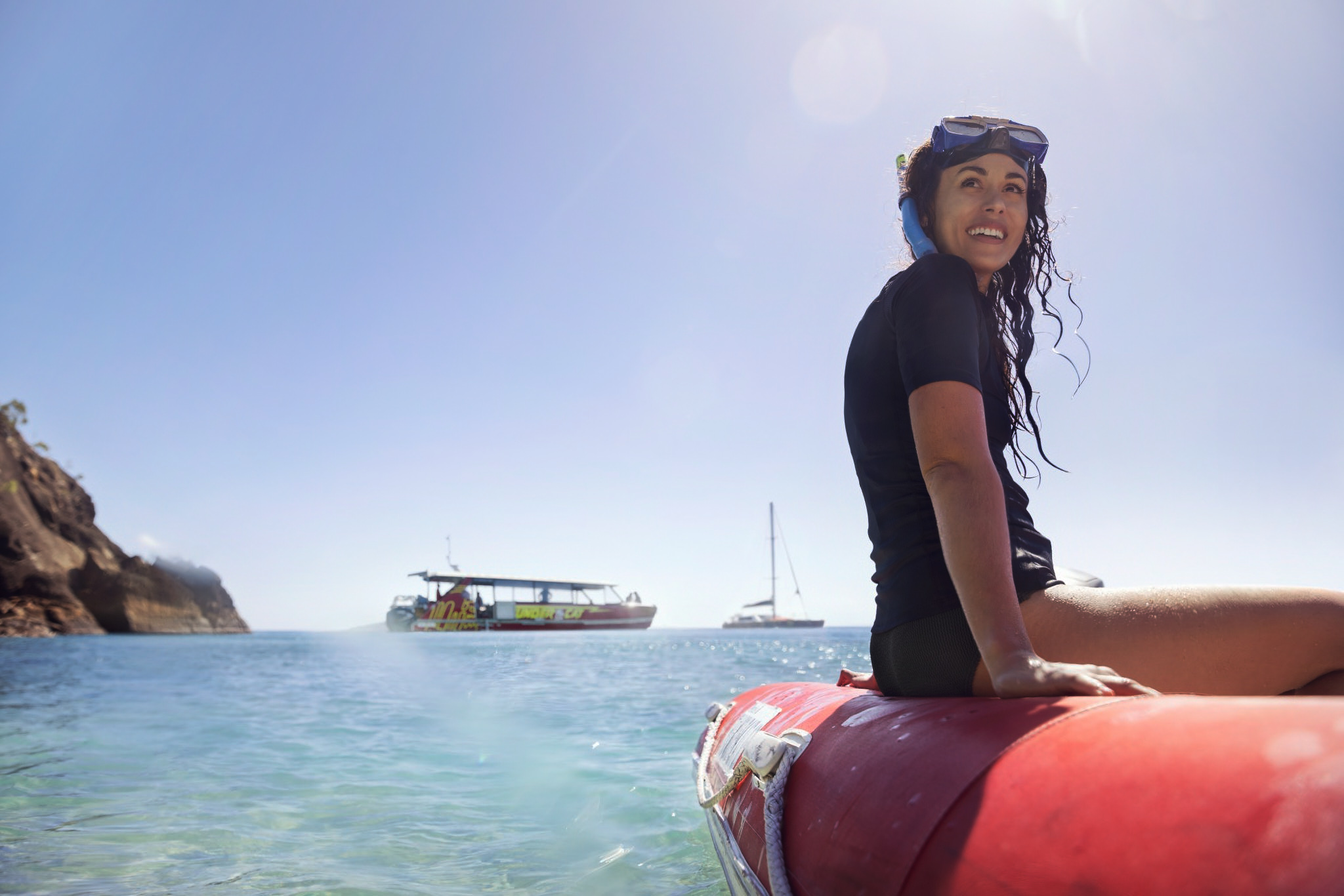 Woman in a rash guard shirt and snorkeling goggles sitting on a red inflatable boat at sea, with boats and a rocky coast in the background.