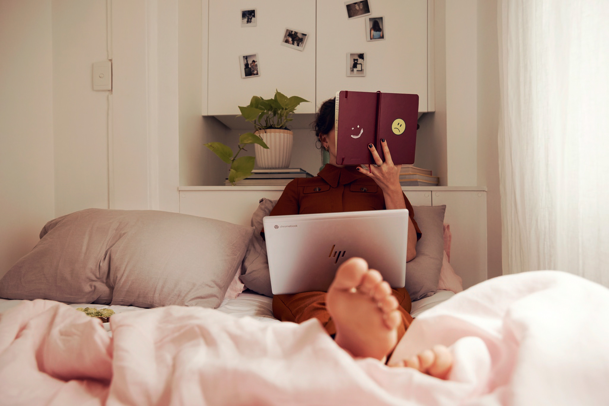 Woman sitting on a bed hiding her face behind a notebook while working on a laptop computer in a bedroom.