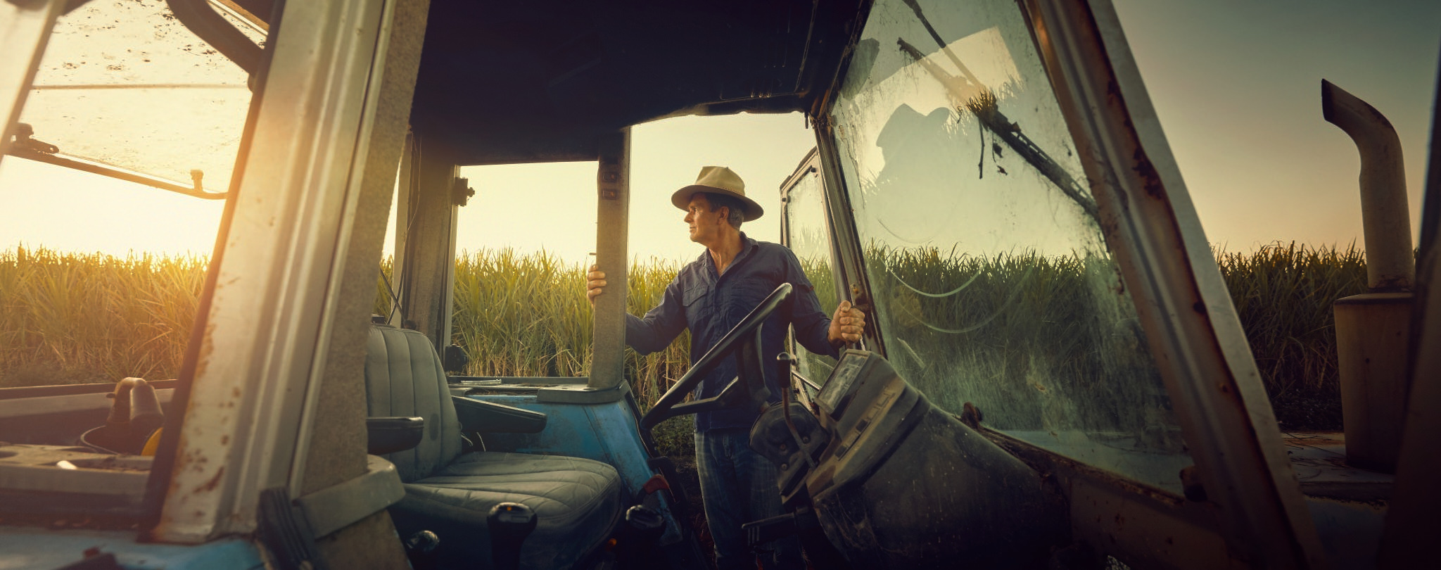 A medium shot shows a male farm worker wearing a hat and blue collared shirt operating a blue tractor during the day in a field.