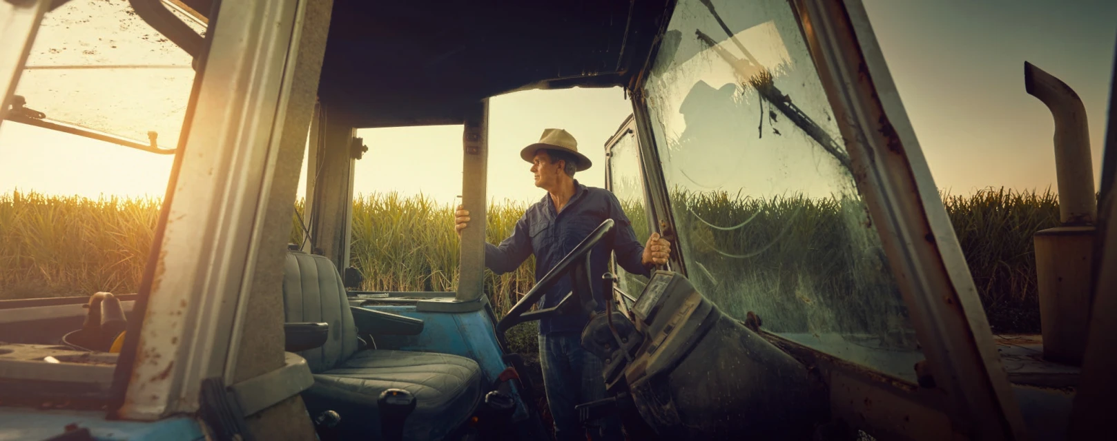 A medium shot shows a male farm worker wearing a hat and blue collared shirt operating a blue tractor during the day in a field.