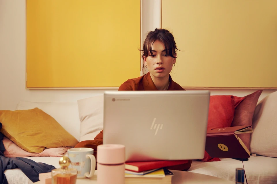 Young adult female works on a laptop while sitting on a couch in a living room with a yellow painting, books, and other objects.