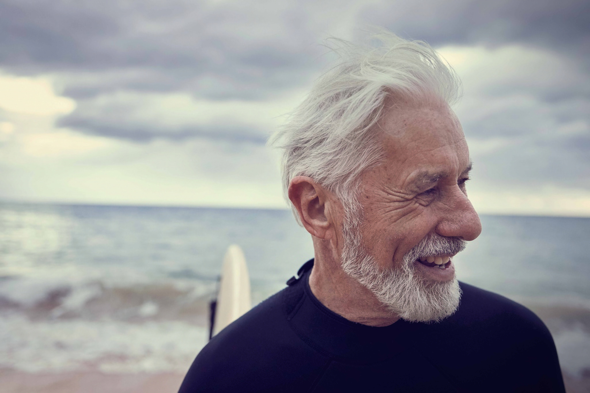 Elderly man with white hair and beard smiling in a wetsuit on a beach with a surfboard