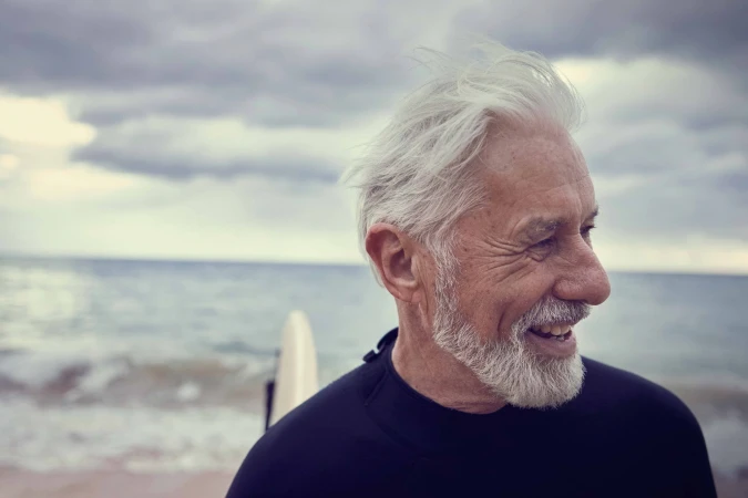 Elderly man with white hair and beard smiling in a wetsuit on a beach with a surfboard