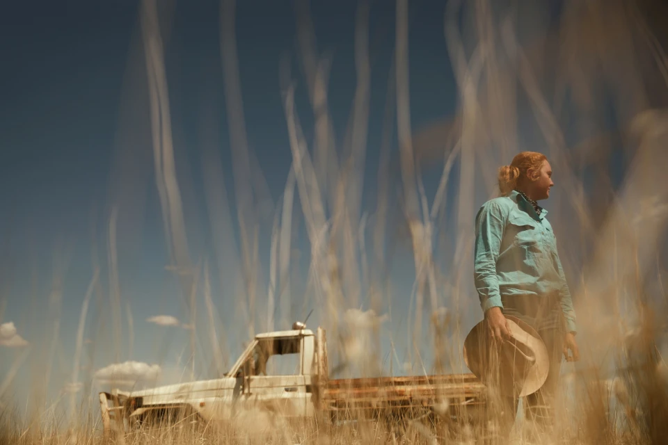 Outdoor portrait of a fair-skinned adult with reddish hair holding a hat, standing in tall grass near a vintage white pickup truck.