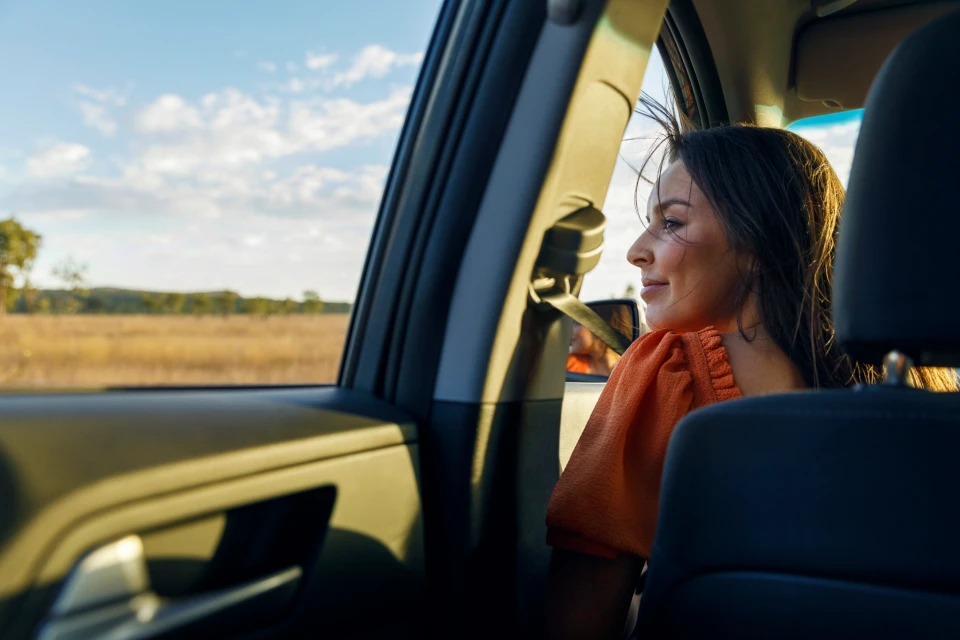 Woman in orange top looks out of car window at sunny, golden field and sky, creating a travel or lifestyle moment.