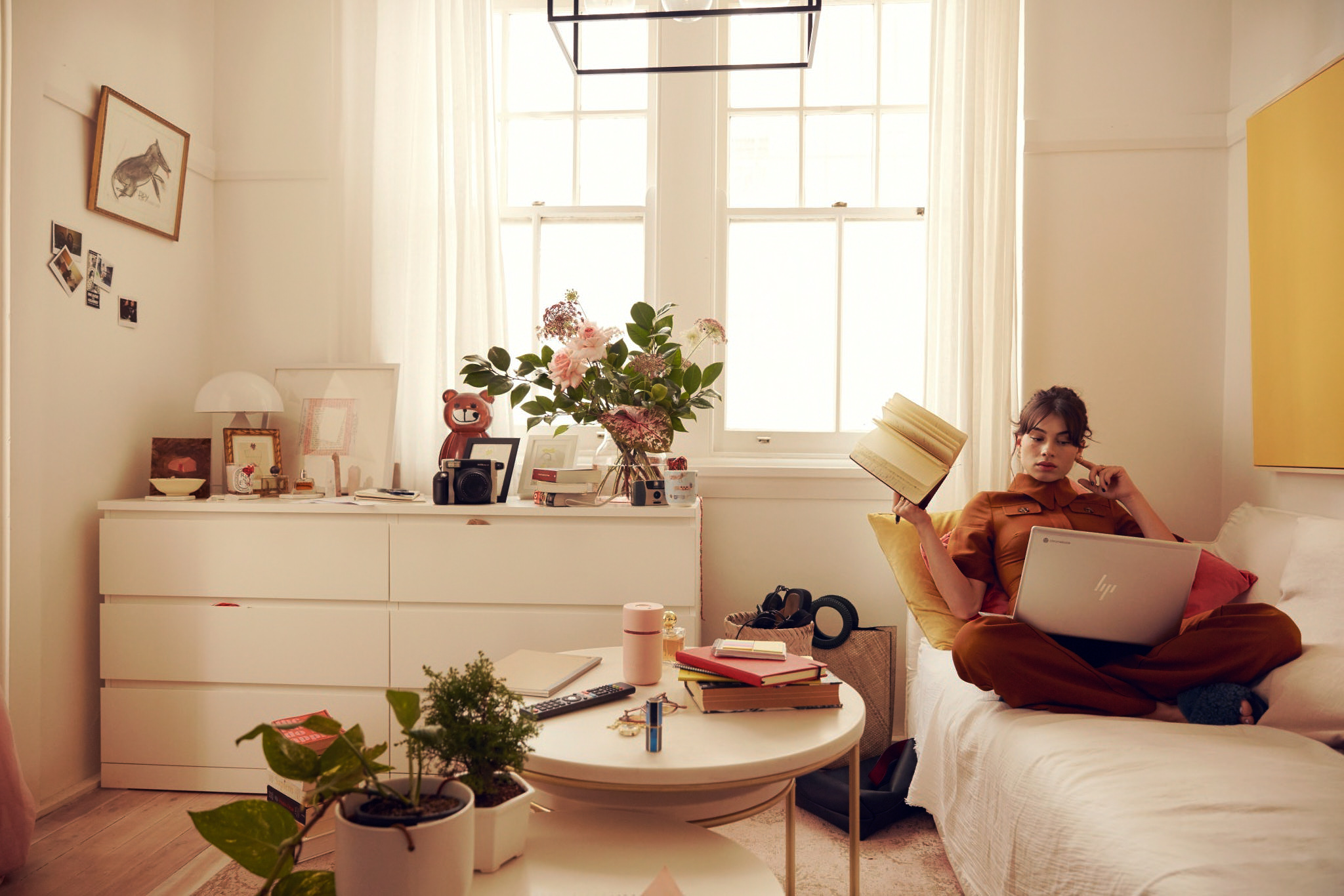 Woman sits on a sofa at home with a laptop, holding a book, surrounded by natural light, plants, and neutral tones in a comfortable setting.