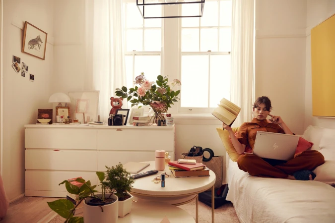 Woman sits on a sofa at home with a laptop, holding a book, surrounded by natural light, plants, and neutral tones in a comfortable setting.