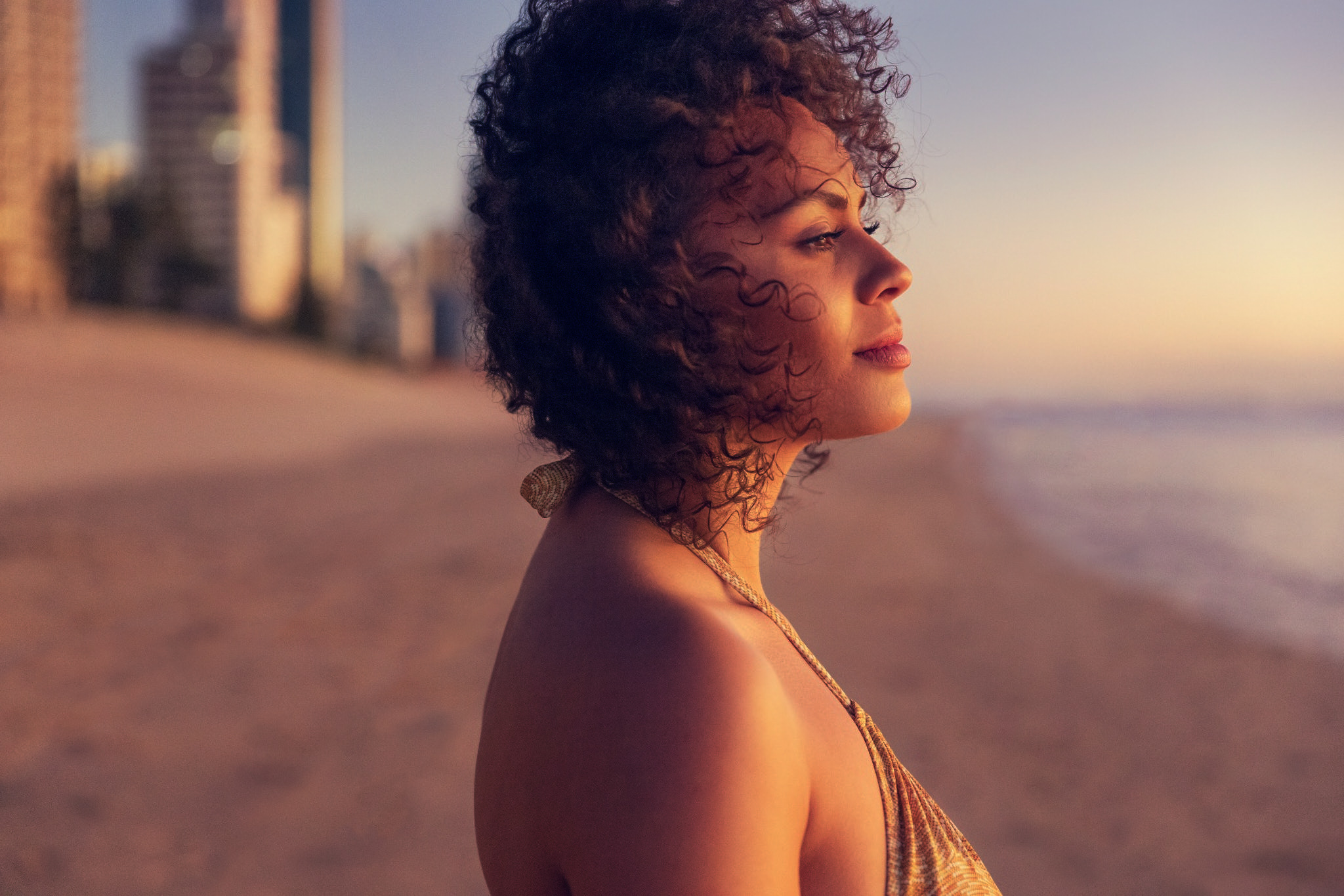 Adult woman with curly hair wearing a golden top, on a sandy beach near the ocean and buildings during sunset. Contemplative facial expression.