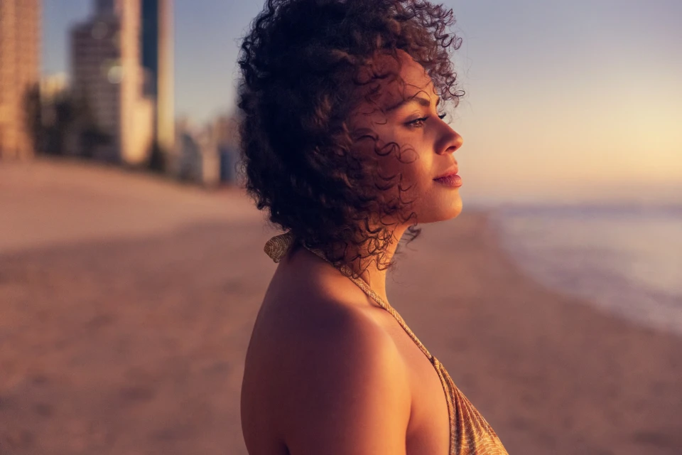 Adult woman with curly hair wearing a golden top, on a sandy beach near the ocean and buildings during sunset. Contemplative facial expression.