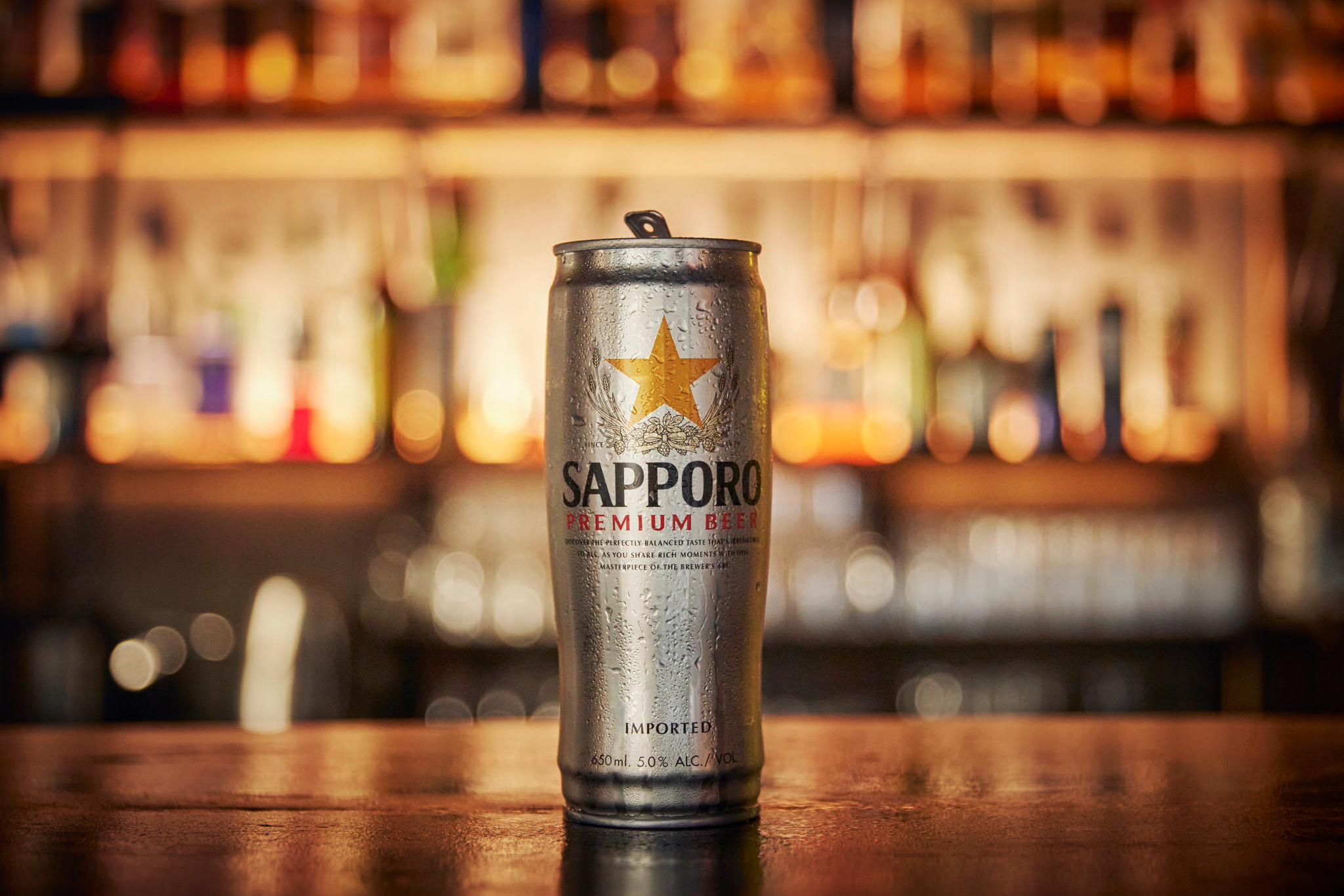 Silver Sapporo Premium Beer can on a wooden bar top with a blurred background featuring lights and bottles in an indoor setting.