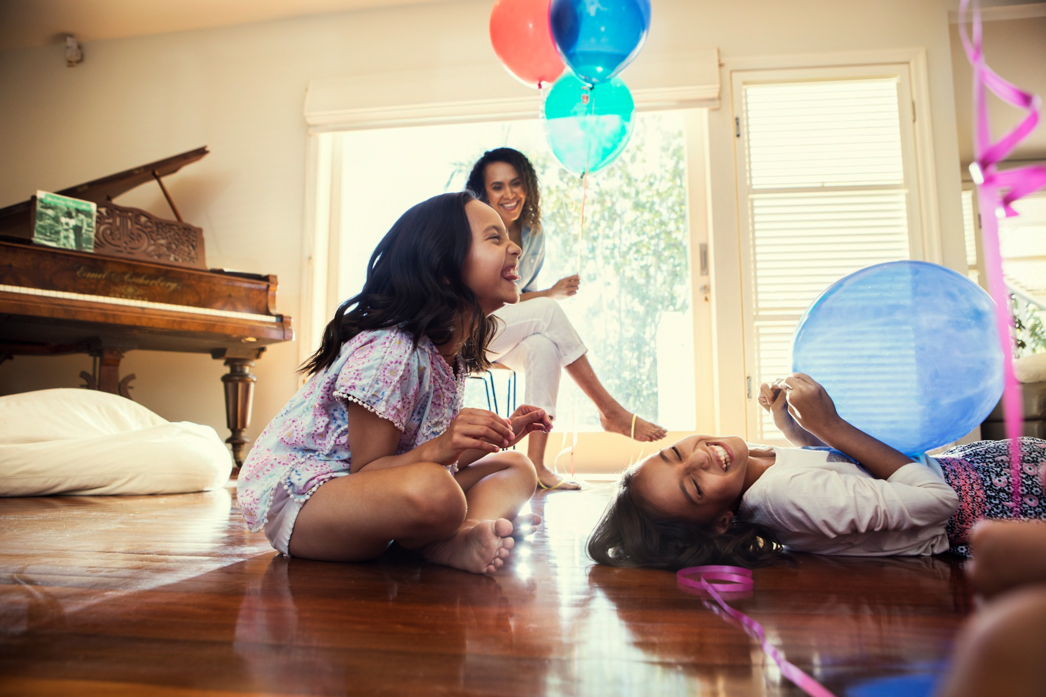 Two smiling girls play on a hardwood floor with balloons, while a woman sits in the background, creating a warm and playful indoor scene.