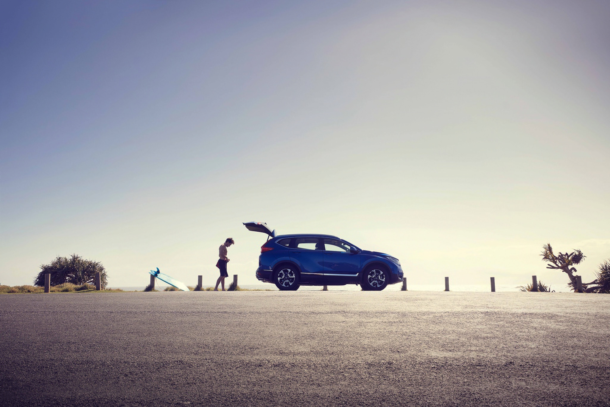 A blue SUV parked near a beach with a surfer and surfboard in the distance under a clear sky on a bright, sunny day.