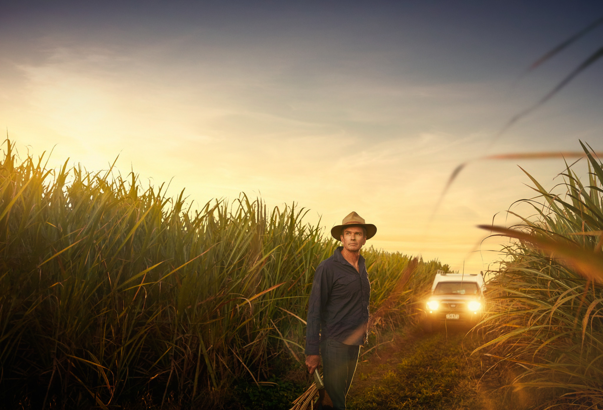 Adult male in a hat stands on a rural farm road amidst tall grasses, with a white truck in the background against a golden sunset.