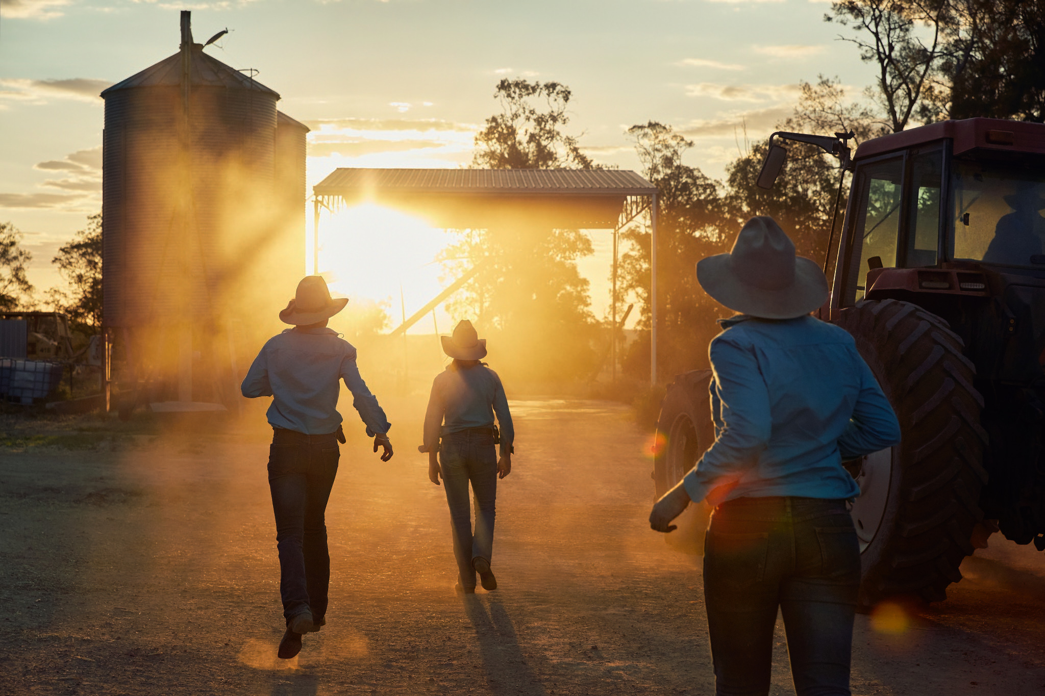 Three adults in work shirts and hats walking toward a tractor and grain silo in a rural setting at sunset with golden sunlight.