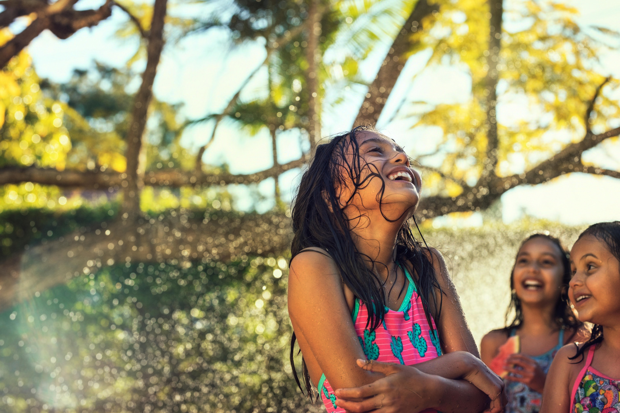 A young girl with wet hair is laughing while being sprayed with water outside, with two other girls visible in the background.