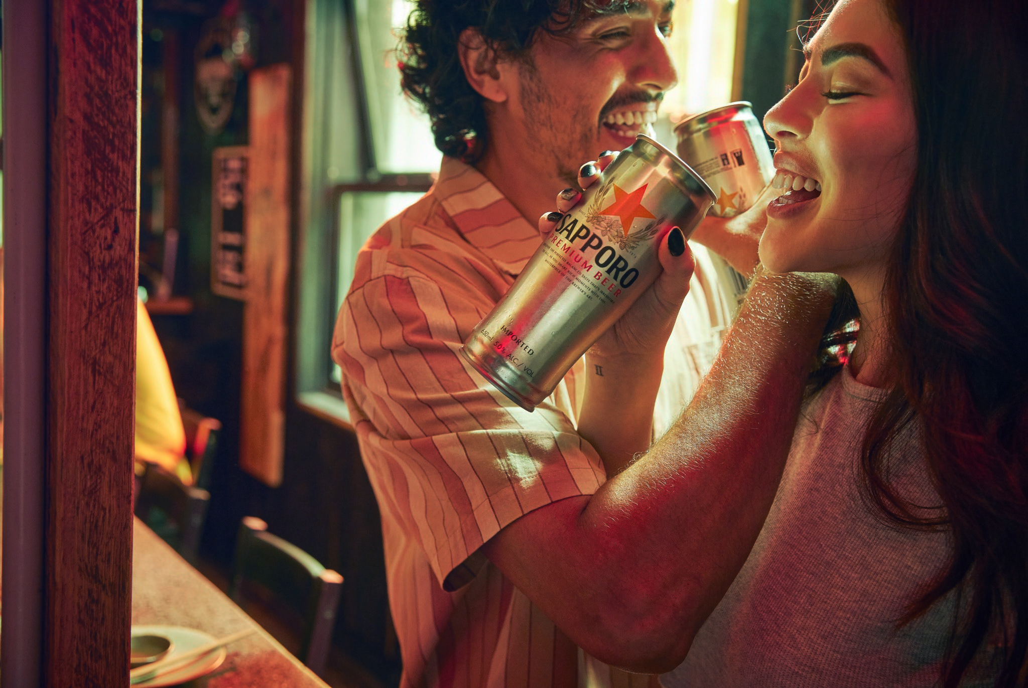 Two adults, a man and a woman, laugh together while holding Sapporo Premium Beer cans in an indoor setting with warm lighting.